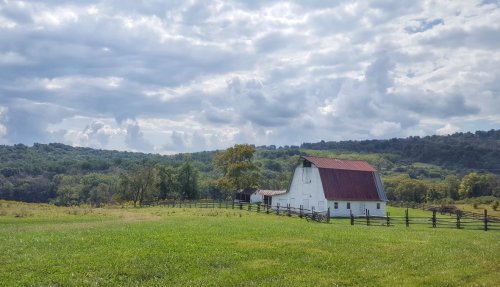 A white barn with a red roof sits in a green field, surrounded by trees and rolling hills under a cloudy sky.