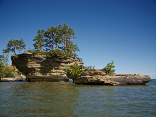 A rocky island with trees stands in calm water under a clear blue sky.