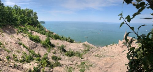 A panoramic view of a sandy cliff overlooking a calm lake with boats and green trees in the background.