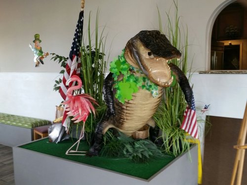 A large alligator statue surrounded by greenery, flags, and a flamingo in a decorative indoor setting.
