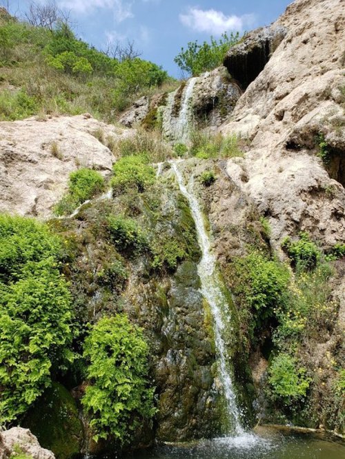 A small waterfall cascading down rocky terrain, surrounded by lush green vegetation under a blue sky.