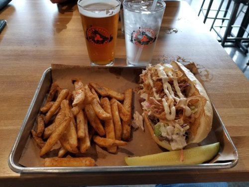 A tray with a sandwich topped with pulled pork, coleslaw, and sauce, served with fries, a pickle, and two drinks.