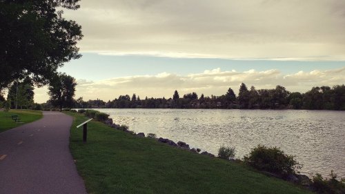A serene riverside path lined with trees, benches, and a calm waterway under a cloudy sky.