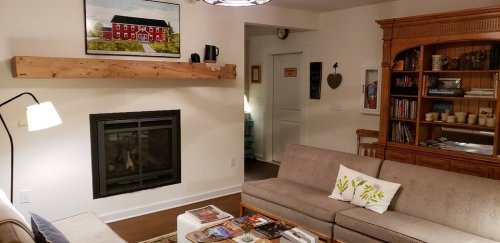 Cozy living room with a fireplace, two sofas, a wooden shelf, and a painting of a red house on the wall.