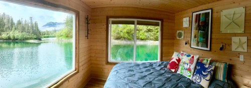 Cozy wooden room with a bed, large window overlooking a serene lake and lush greenery. Decorated with colorful pillows.