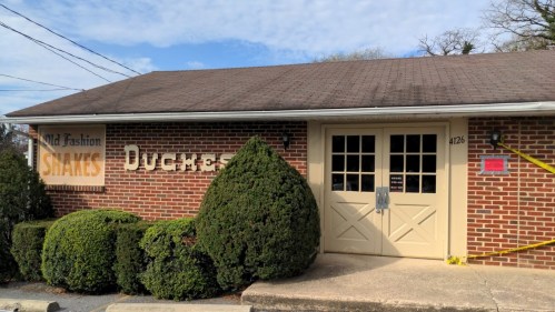 A brick building with a sign reading "Old Fashion Shakes" and "Duchess" near neatly trimmed bushes and a cloudy sky.