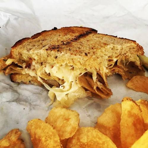 A half-eaten sandwich on toasted bread with layers of filling, accompanied by a side of potato chips.