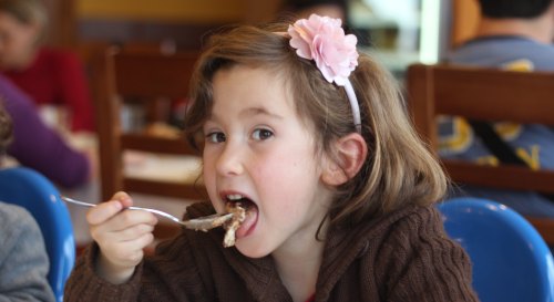 A young girl with a flower headband happily eating with a fork in a restaurant setting.