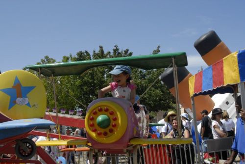 A joyful child in a blue hat rides a colorful airplane at a fair, surrounded by people and festive decorations.