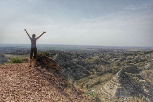 A person stands on a rocky ledge, arms raised, overlooking a vast, rugged landscape under a hazy sky.