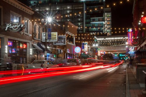 A vibrant city street at night, featuring illuminated signs, bustling traffic, and string lights overhead.