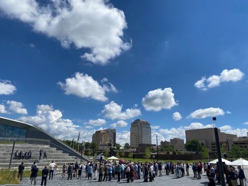 A crowd gathers outdoors under a blue sky with fluffy clouds, surrounded by modern buildings and greenery.