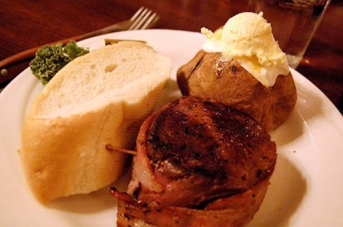 A plate featuring a grilled steak wrapped in bacon, a baked potato with butter, a slice of bread, and a garnish of lettuce.