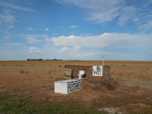 A rustic outdoor setup featuring a toilet and a chair, with graffiti reading "REST AREA" and "WIFI," against a vast field.