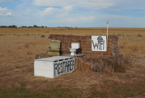 A quirky outdoor setup with a toilet, chair, and signs for "WiFi" and "Rest Area" on a hay bale backdrop.