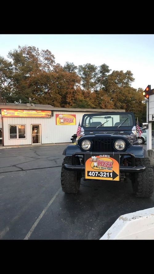 A blue jeep with flags parked in front of a pizza shop, featuring a colorful sign with a phone number.