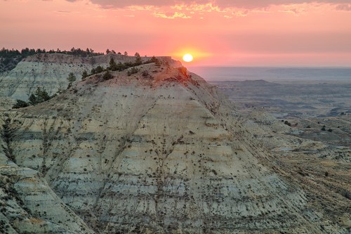 Sunset over layered hills in a vast landscape, with soft clouds and a warm glow illuminating the scene.