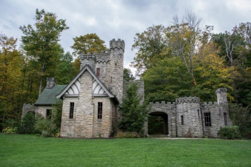 A stone castle with turrets surrounded by trees and greenery under a cloudy sky.