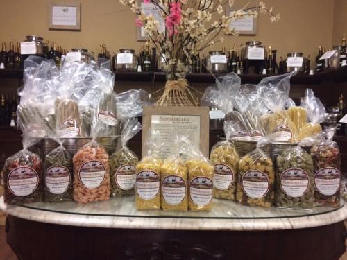 A display of various pasta types in clear bags, arranged on a marble counter with decorative items in the background.
