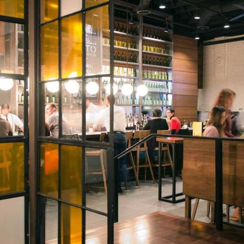A lively restaurant interior with a bar, patrons socializing, and shelves filled with bottles in the background.