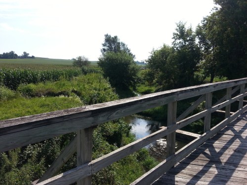 A wooden bridge spans a small creek, surrounded by lush greenery and fields under a clear blue sky.