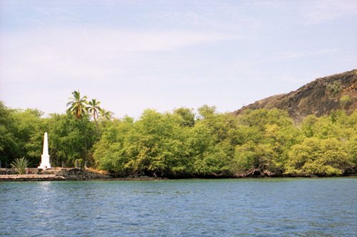 A serene landscape featuring a monument by the water, surrounded by lush greenery and a rocky hillside in the background.