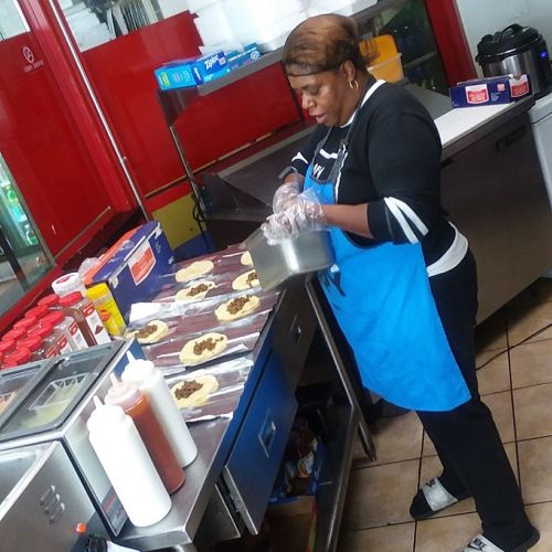 A woman in an apron prepares food at a counter, surrounded by condiments and cooking supplies in a restaurant kitchen.