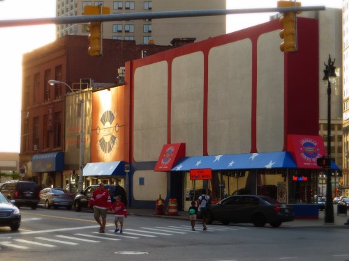 A street view of a colorful building with red and blue awnings, pedestrians crossing, and cars on the road.