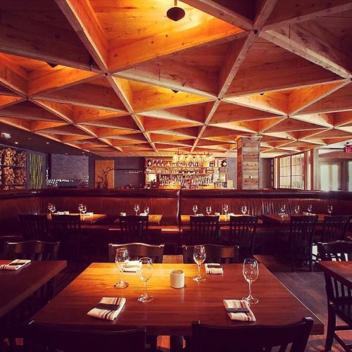 Interior of a modern restaurant featuring wooden ceiling design, cozy seating, and a well-stocked bar in the background.