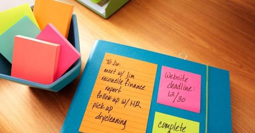 A blue folder with sticky notes listing tasks, alongside a bowl of colorful sticky notes on a wooden desk.
