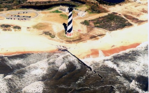 Aerial view of a striped lighthouse on a sandy beach, with waves crashing and a parking area nearby.