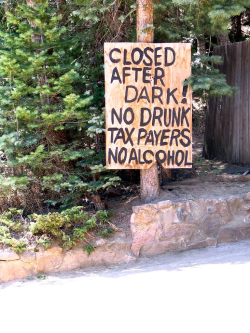 A wooden sign on a tree reads: "CLOSED AFTER DARK! NO DRUNK TAX PAYERS NO ALCOHOL." Surrounded by greenery.