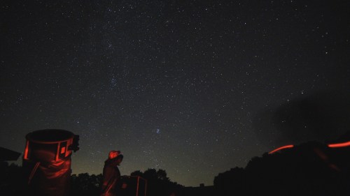 A person stands under a starry night sky, with telescopes and red lights illuminating the scene.