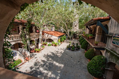 A charming courtyard with stone pathways, lush greenery, and rustic buildings under a clear blue sky.