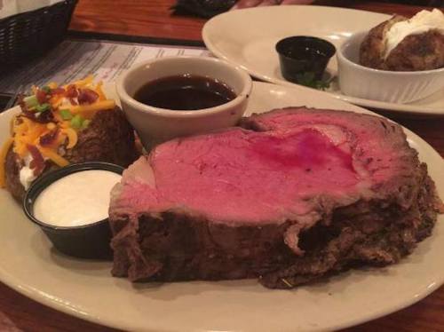 A plate featuring a large slice of rare prime rib, a baked potato with toppings, and dipping sauces.