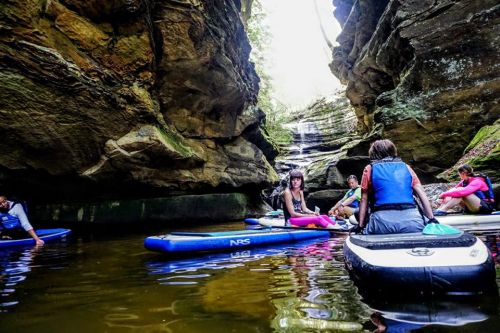 Group of people paddleboarding in a narrow canyon with a waterfall in the background. Lush greenery surrounds the scene.