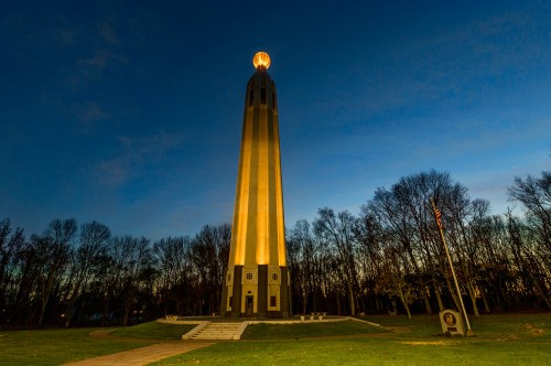 A tall monument illuminated at dusk, surrounded by trees and a grassy area, with a glowing orb at the top.