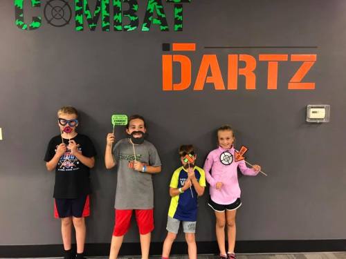 Four kids holding fun props in front of a "COMBAT DARTZ" wall sign, smiling and posing for the camera.