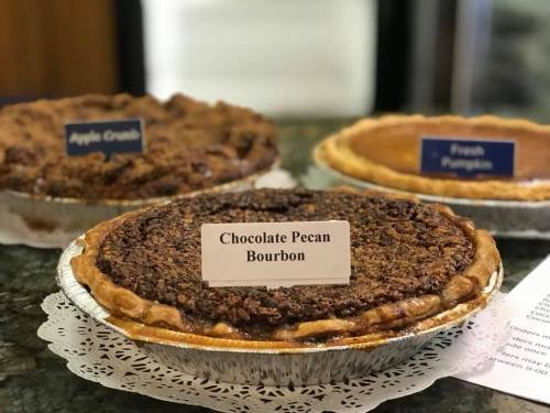 Three pies on a table: Chocolate Pecan Bourbon, Apple Crumble, and Fresh Pumpkin, each labeled with a sign.