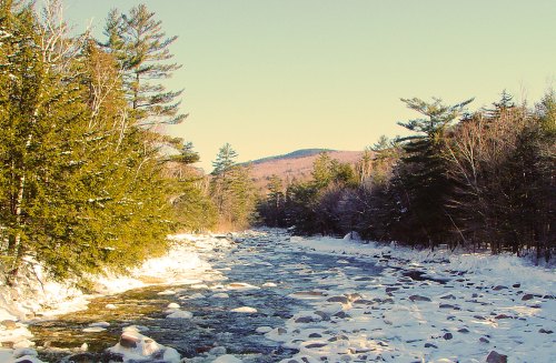 A serene winter landscape featuring a river surrounded by snow-covered rocks and evergreen trees under a clear sky.
