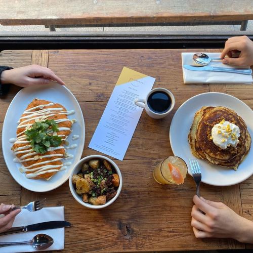 A table with two plates of food: one with pancakes and the other with a savory dish, plus a drink and a menu.
