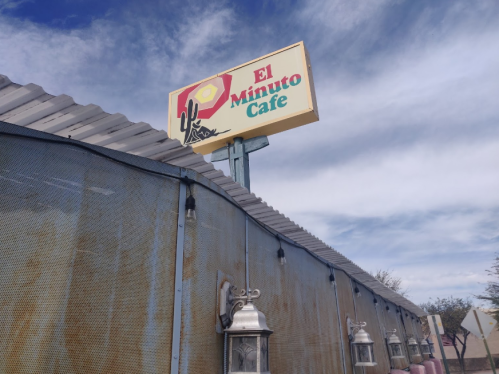 Sign for El Minuto Cafe above a rustic building with a metal roof and lanterns, under a blue sky with clouds.