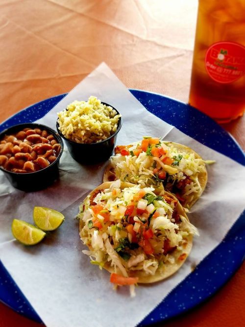 Two tacos filled with fresh toppings, served with rice, beans, and lime wedges on a blue plate. Iced tea in the background.