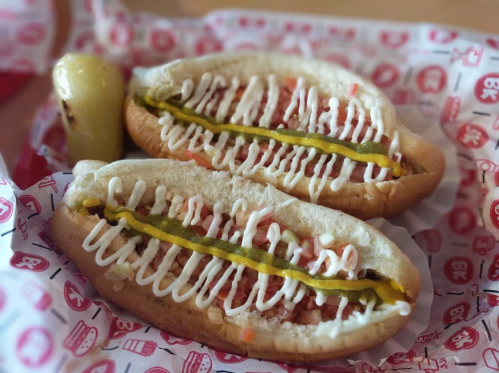 Two hot dogs in a bun topped with mayonnaise, mustard, and diced tomatoes, served in a red and white paper basket.