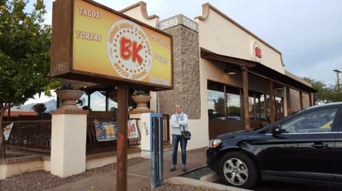 A woman stands outside a restaurant named "BK," with a sign displaying tacos, tortas, and hot dogs. A car is parked nearby.