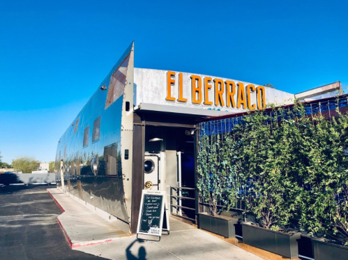 Exterior of El Berraco restaurant featuring a shiny metal facade and vibrant blue accents under a clear blue sky.