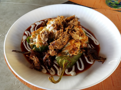 A plate of stuffed poblano peppers topped with shredded meat, cheese, and drizzled with sauce and crema.