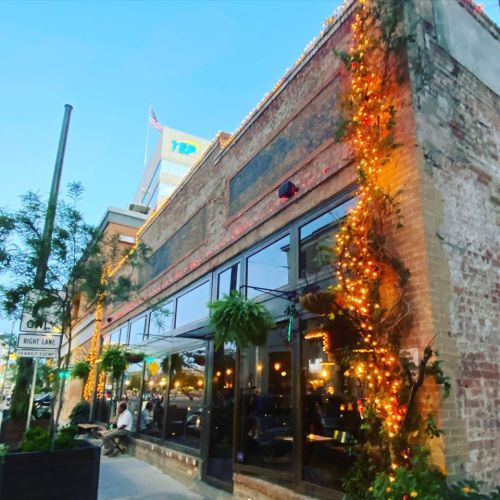 A brick restaurant exterior adorned with string lights and greenery, set against a twilight sky.