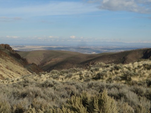A panoramic view of rolling hills and valleys, with sparse vegetation and distant mountains under a clear blue sky.