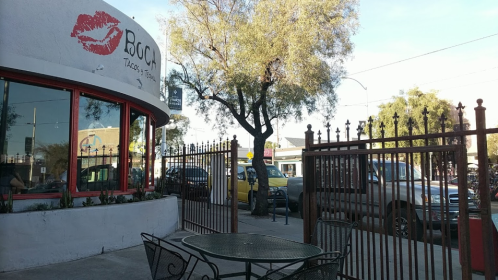 Outdoor seating area at a taco restaurant, with a tree and a view of parked cars in the background.